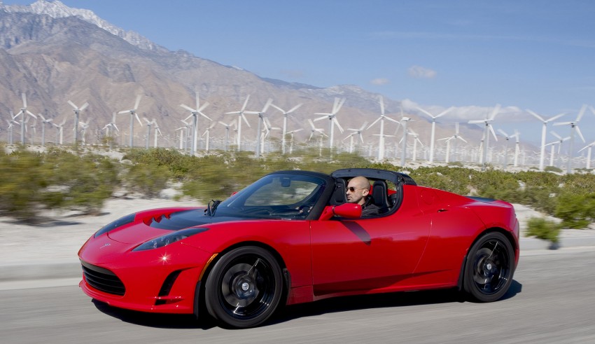 A red tesla roadster driving in front of wind turbines.
