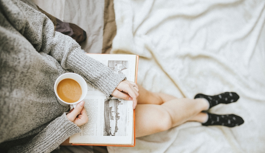 A woman holding a book and a cup of coffee on a bed.