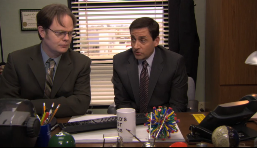 Two men sitting at a desk talking to each other.