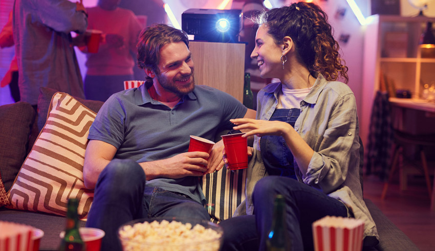 A man and woman sitting on a couch watching tv and drinking popcorn.