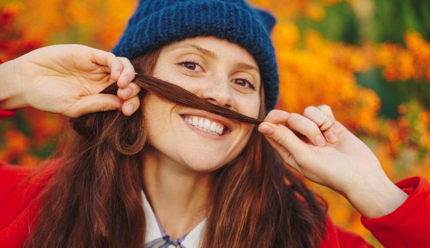 A woman with a red beanie is putting her finger on her mustache.