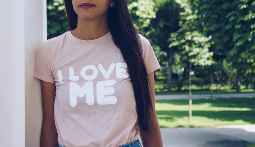 A woman wearing a pink i love me t - shirt leaning against a wall.