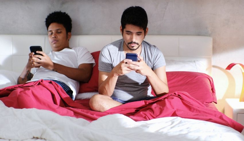 Two young men sitting on a bed looking at their cell phones.