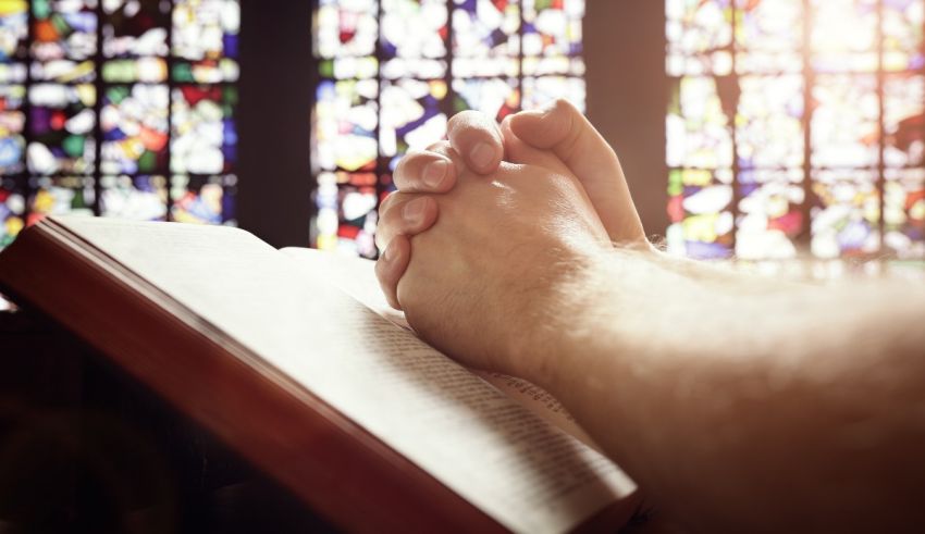 A man's hands are resting on a bible in front of stained glass windows.