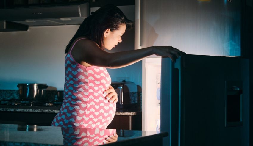 A pregnant woman looking at the refrigerator at night.