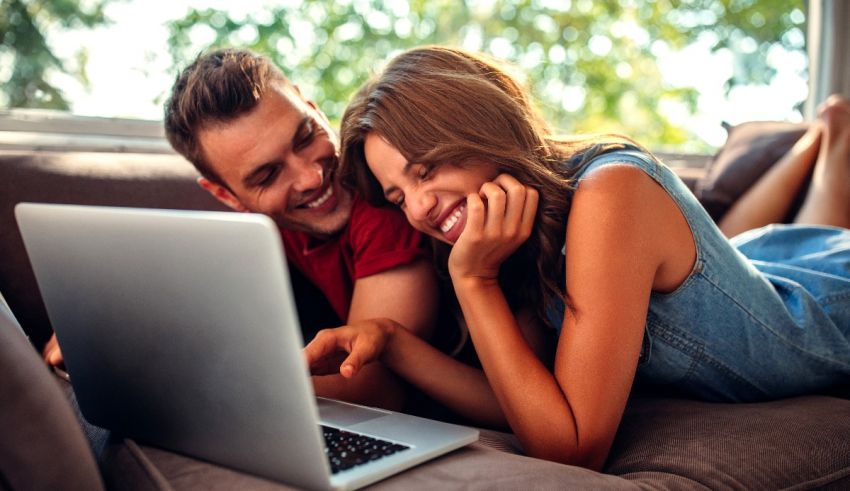 A man and woman are laying on a couch and using a laptop.