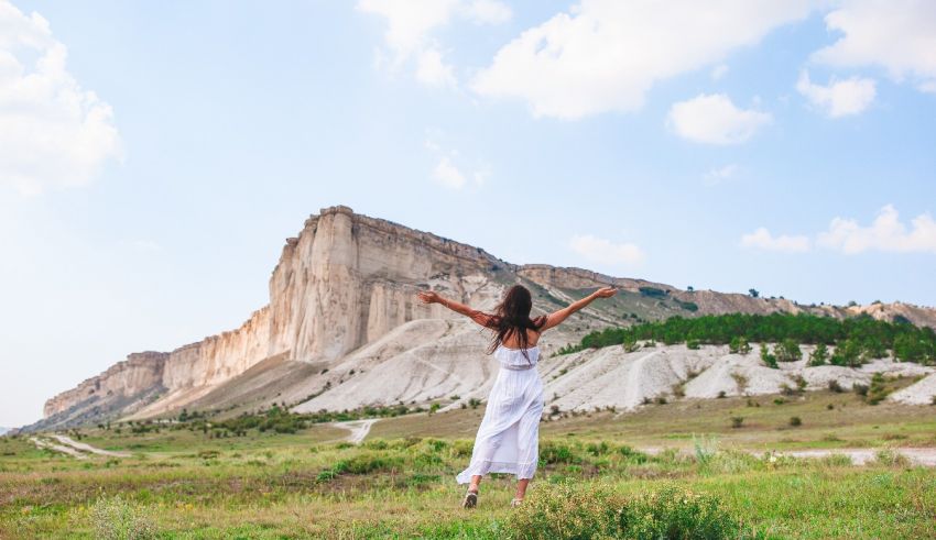 A woman in a white dress standing in front of a mountain with her arms outstretched.