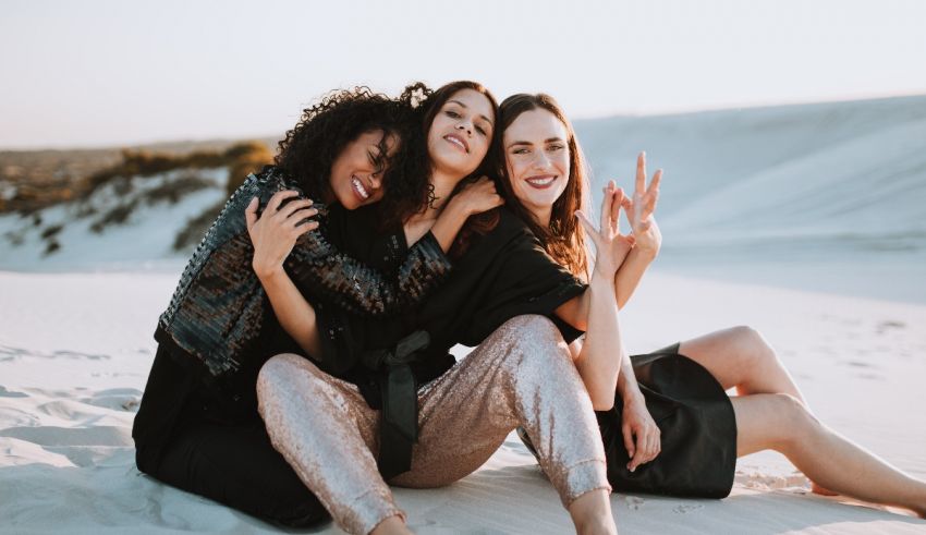 Three women sitting on a sand dune and making a peace sign.