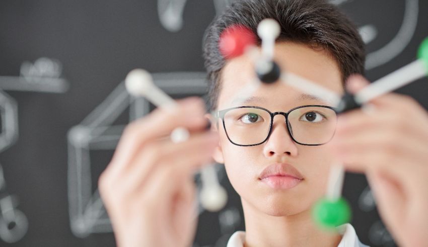 A young boy is holding up a molecule in front of a blackboard.