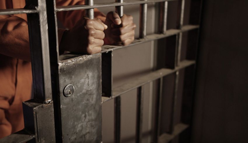A man in an orange prison uniform holding his hands out of a jail cell.