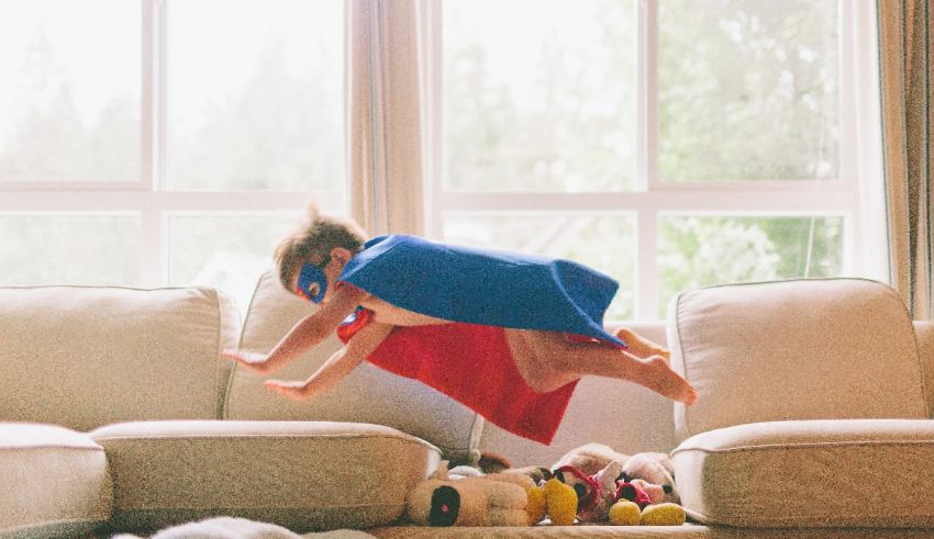 A child in a superhero cape jumping on top of a couch.