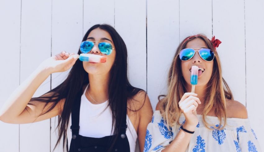 Two young women are eating ice pops in front of a white wall.