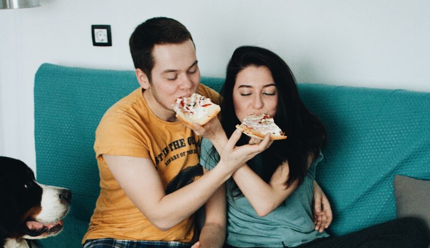 A man and woman eating pizza on a couch with a dog.