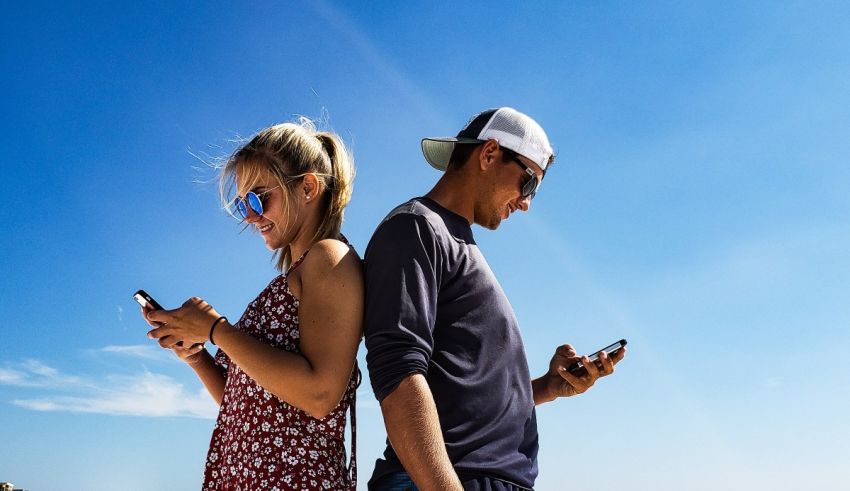 A man and woman standing on a beach looking at their cell phones.