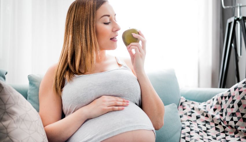 A pregnant woman eating an apple while sitting on a couch.