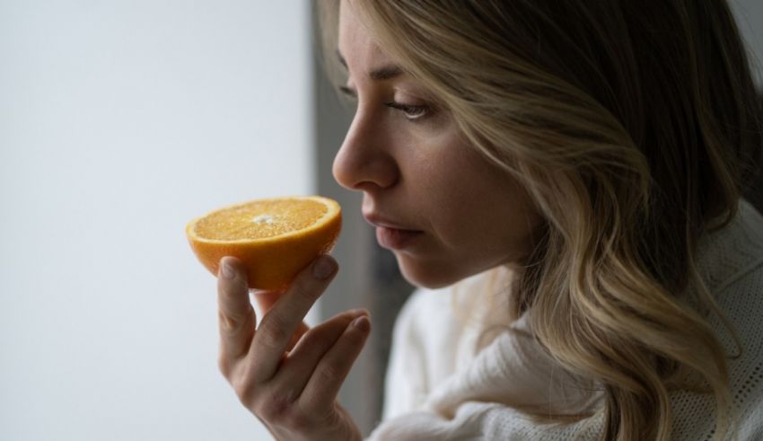 A woman is smelling an orange in front of a window.