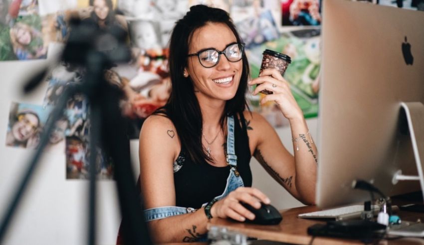 A woman smiling at her computer while holding a cell phone.