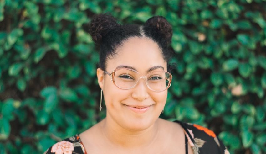 A woman with glasses smiling in front of a bush.