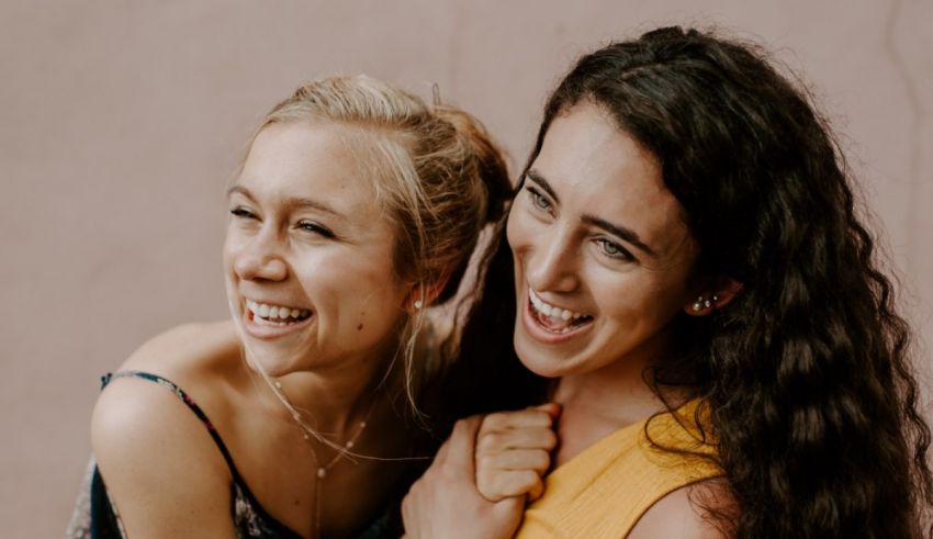 Two young women laughing together in front of a pink wall.
