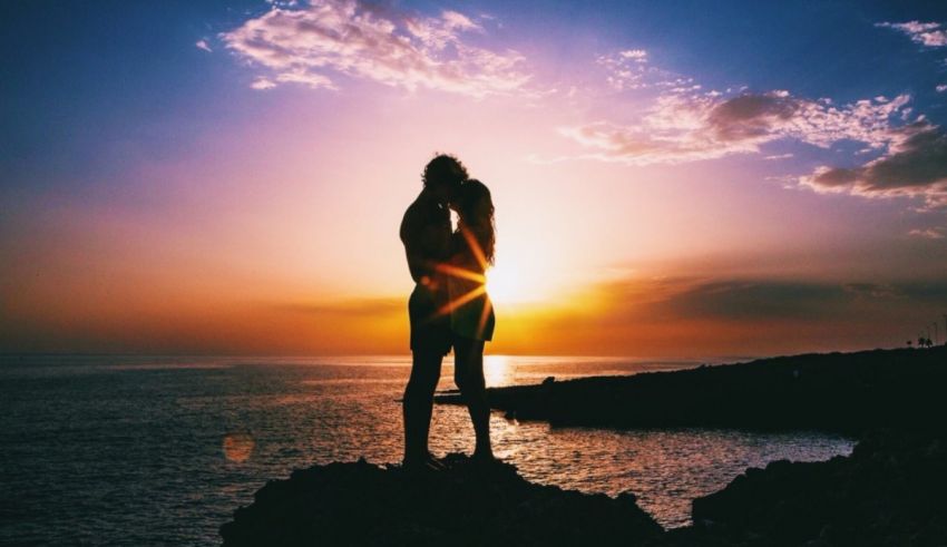 A couple is standing on top of a cliff with the sun setting behind them.