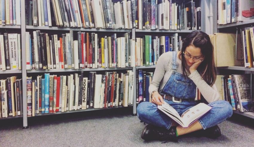 A girl sitting on the floor reading a book in a library.