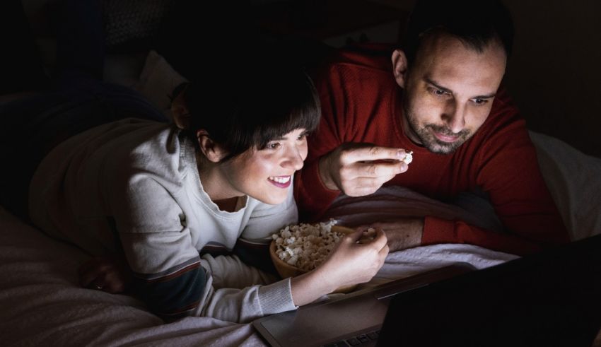 A man and woman laying on a bed with popcorn and a laptop.