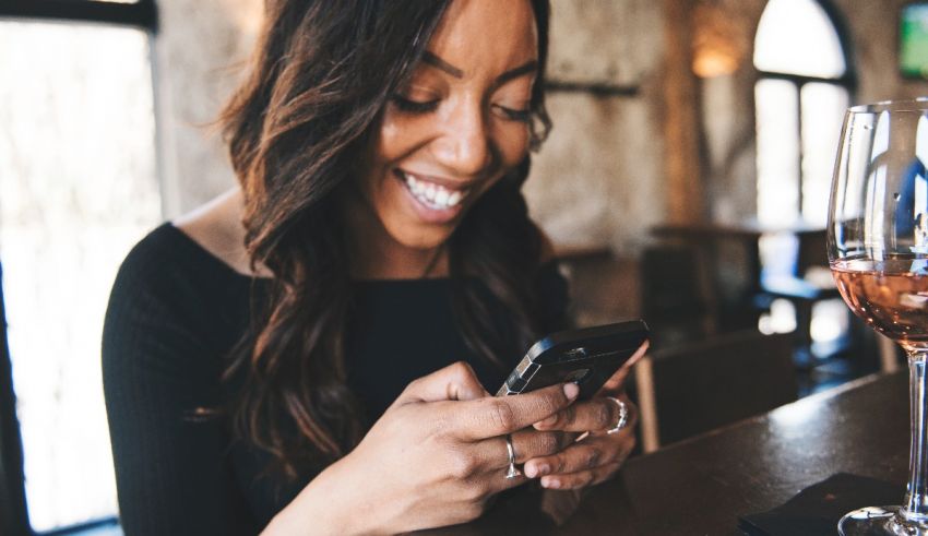 A woman smiling at her phone while holding a glass of wine.