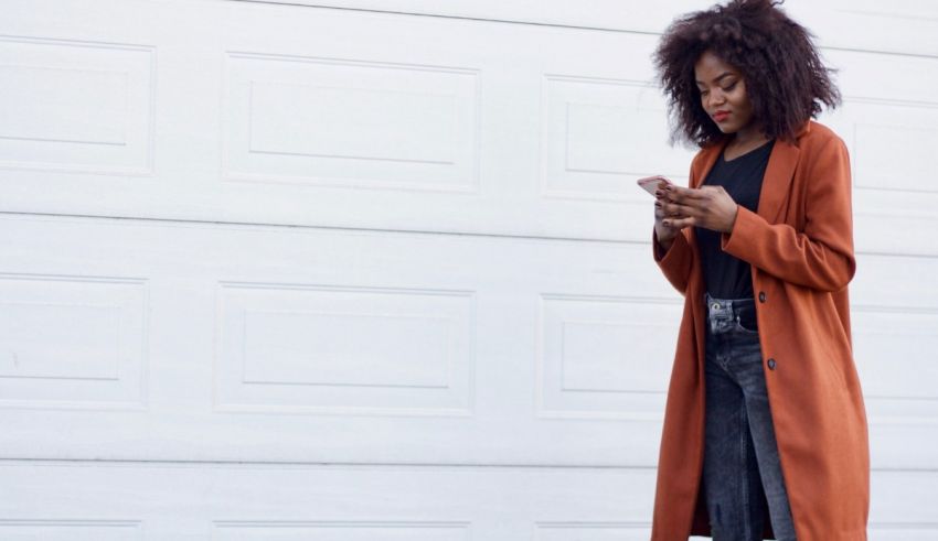 A black woman in an orange coat is using her cell phone in front of a garage.