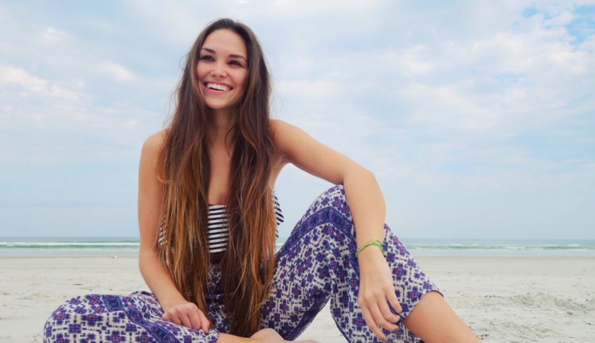 A young woman sitting on the beach in floral pants.