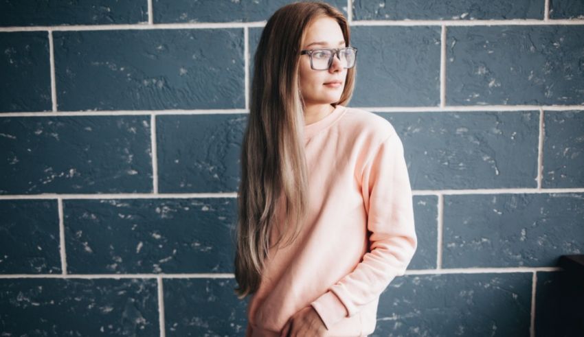 A young woman wearing glasses and a pink sweater leaning against a blue wall.