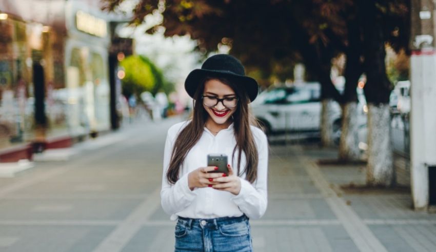 A young woman wearing a hat and glasses is looking at her phone.