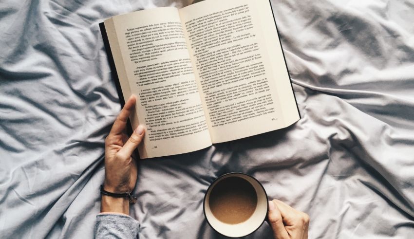 A person reading a book on a bed with a cup of coffee.