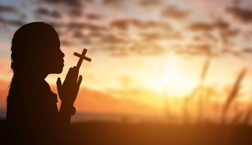 A silhouette of a woman praying with a cross at sunset.