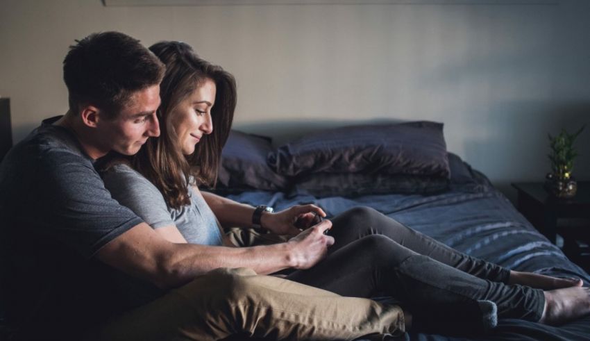 A man and woman sitting on a bed looking at a cell phone.