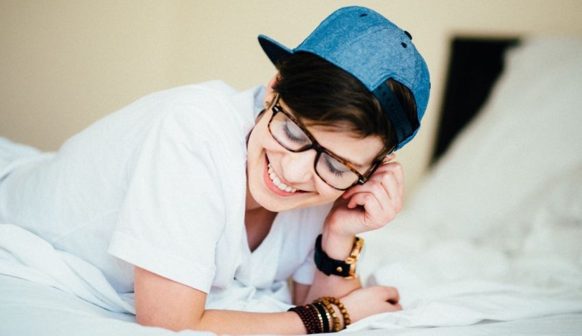 A young man wearing glasses and a hat laying on a bed.