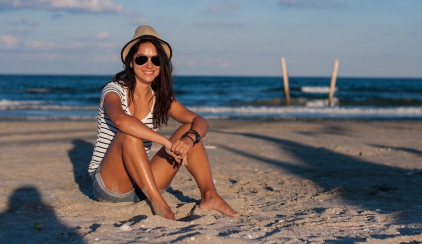 A young woman sitting on the beach wearing a hat and sunglasses.
