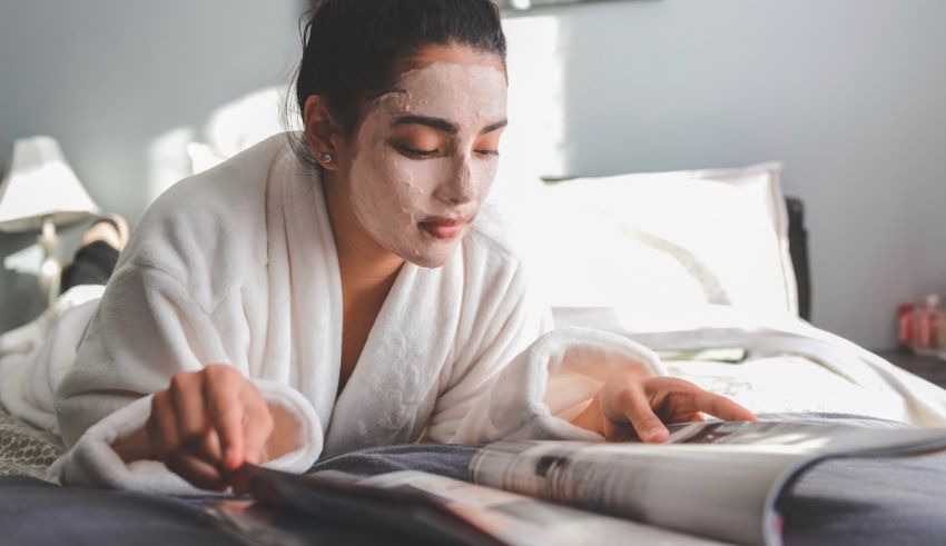 A woman in a robe reading a magazine while wearing a facial mask.