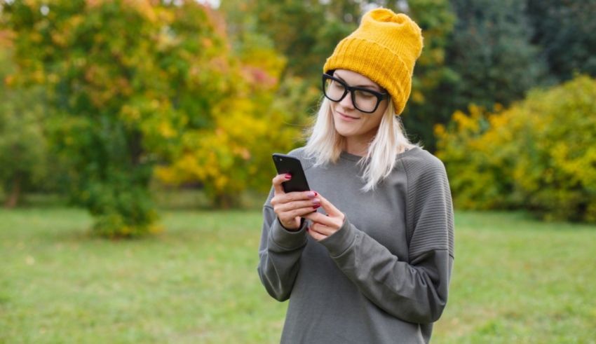 A young woman wearing a yellow beanie and glasses is using her phone in a park.