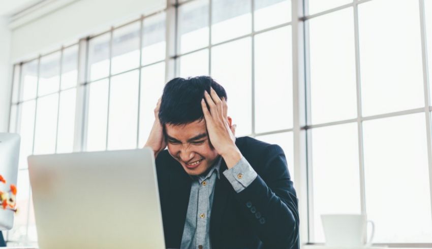 A man in a suit is holding his head while working on a laptop.