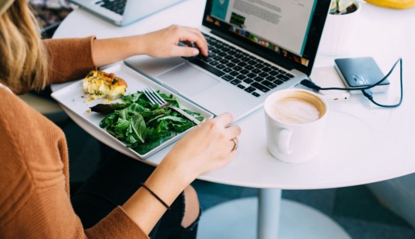 A woman working on her laptop at a table with a salad and a cup of coffee.