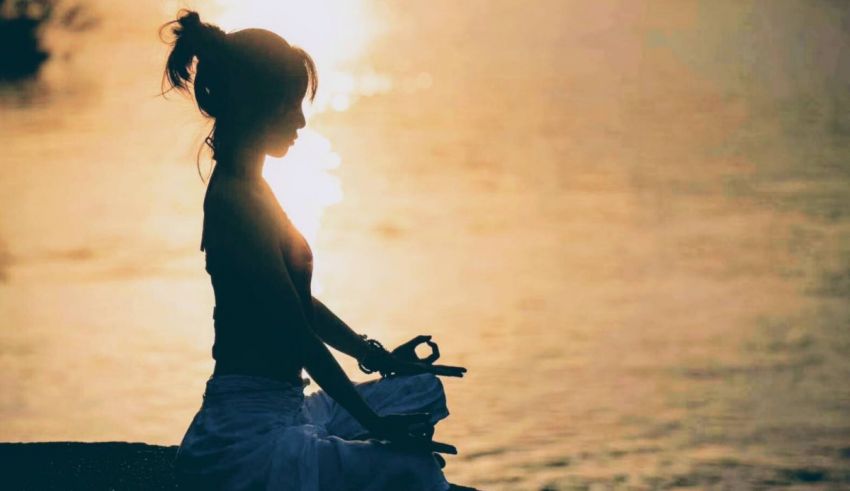 A woman is meditating by the water at sunset.
