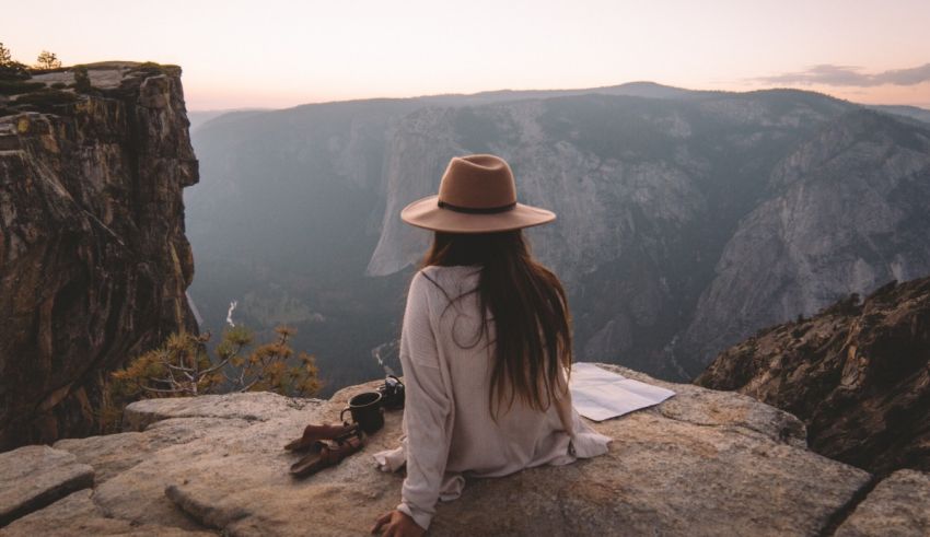 A woman in a hat sits on a cliff overlooking yosemite national park.
