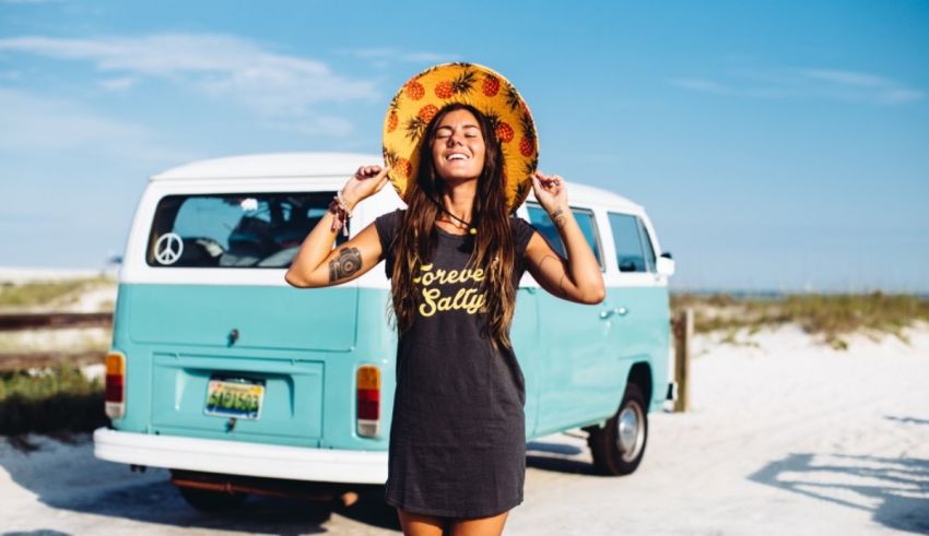 A woman wearing a hat standing next to a vw van on the beach.
