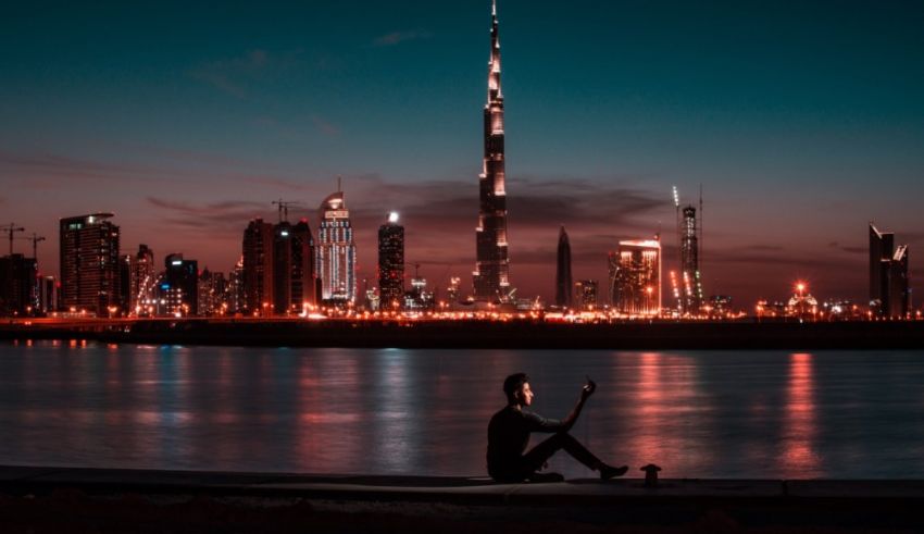 A man sits on the edge of the water at dubai skyline at night.