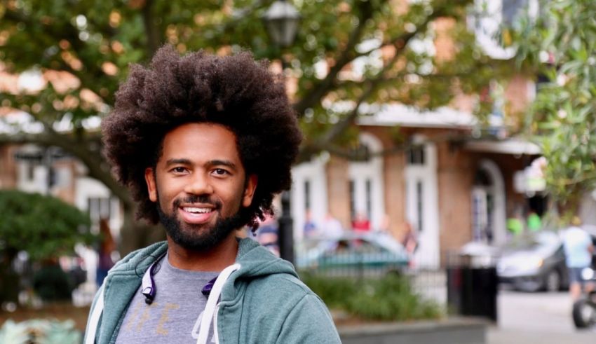 A man with an afro smiles in front of a building.
