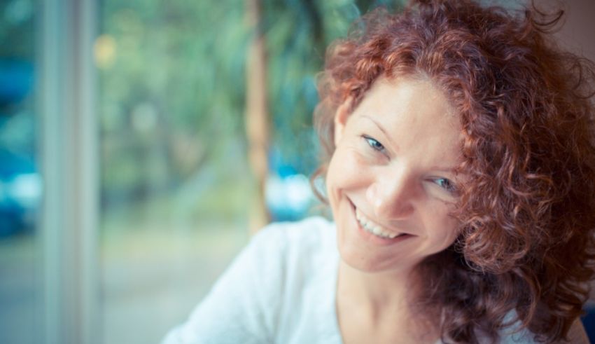 A woman with red curly hair smiling in front of a window.