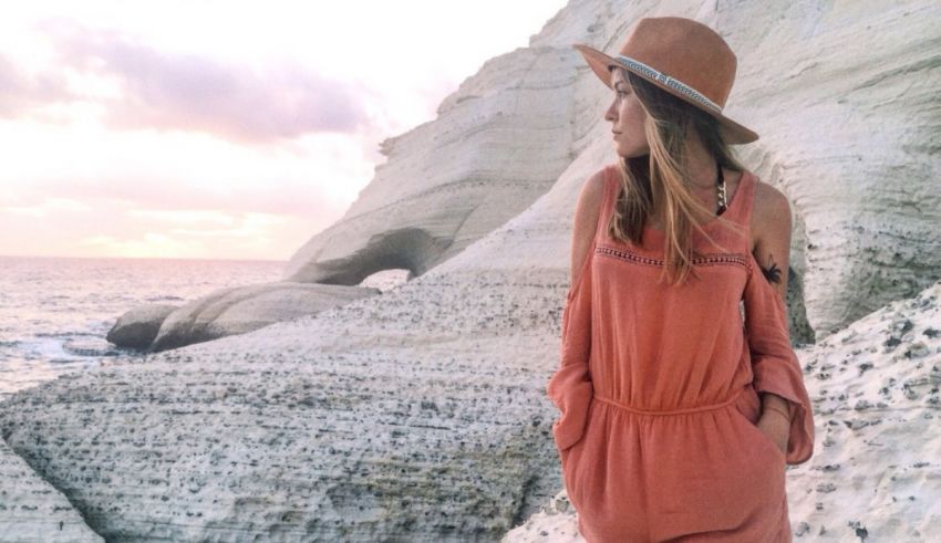 A woman wearing a pink dress and hat is standing on a rocky beach.