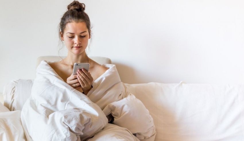 A woman sitting on a couch covered in a blanket looking at her phone.