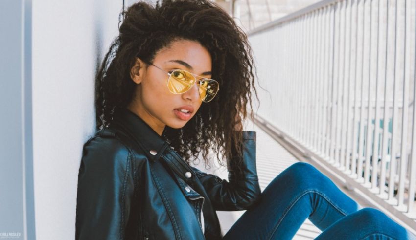 A black woman wearing sunglasses and a leather jacket leaning against a wall.
