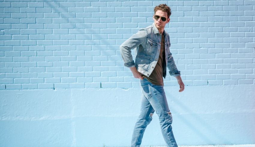 A man in jeans and a denim jacket walking in front of a blue wall.